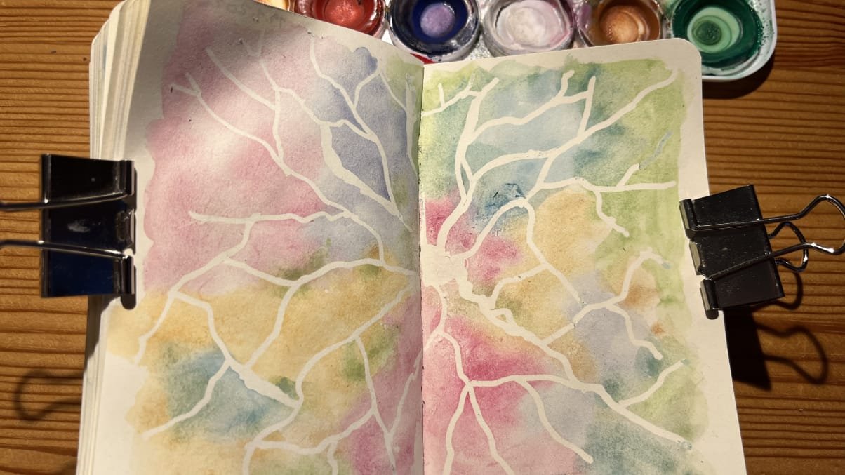 Photo of an open sketchbook on a wooden table. The sketchbook spread is filled with colourful pastelly watercolour and white network like lines spreading from the middle to the sides. On the topside of the sketchbook there's a partial watercolour palette visible.