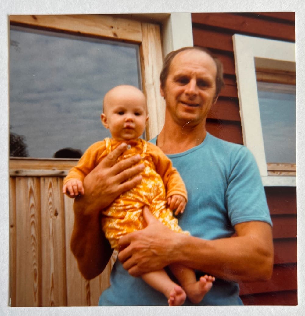 An old photo of my dad holding my as a baby in front of a door of a wooden house. Dad is a balding guy wearing a blue t-shirt and I'm bald baby, wearing a yellow shirt and yellow overalls with some kind of a white pattern, maybe flowers. Dad looks like he's saying something and my toes are pointing up.