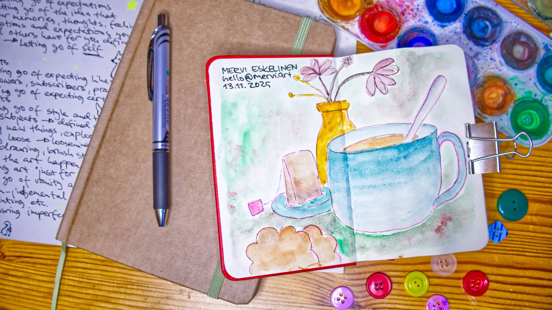 Photo of stuff on wooden table: An open sketchbook filled with a still life painting of tea with cookies and flowers, an open and messy watercolour palette, buttons, fake flowers, a journal and pen and on the bottom an open notebook with partial text on it